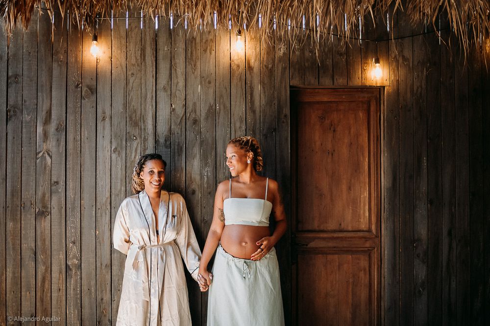 Two women holding hands and smiling together against a rustic wooden wall with warm string lights.