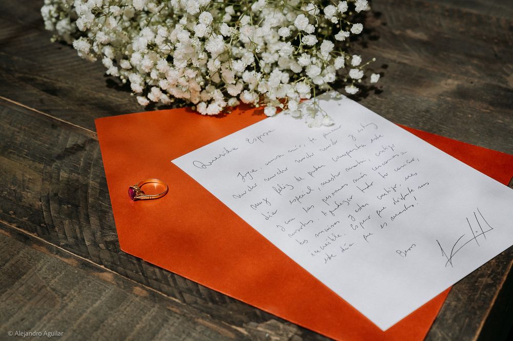 Handwritten wedding letter on white paper with orange envelope, gold ring, and white baby's breath flowers on rustic wood.