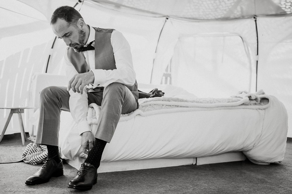 Groom sitting on bed in tent, putting on dress shoes while wearing vest and bow tie before wedding ceremony.
