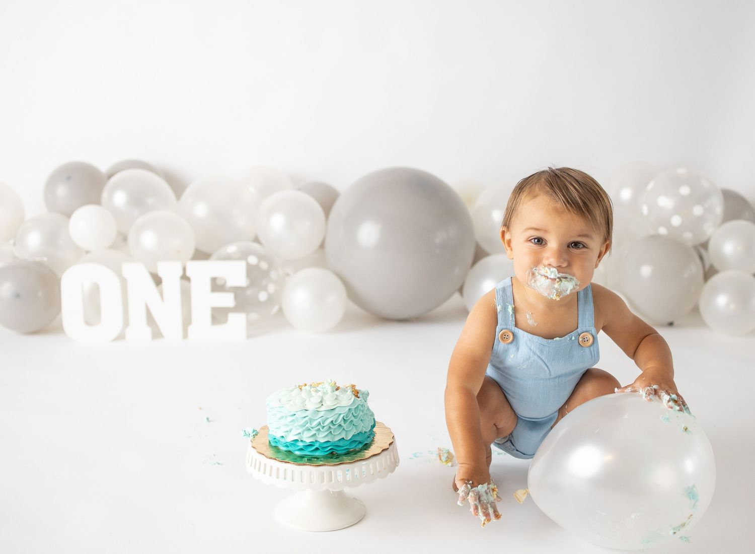 Baby boy in blue romper celebrates first birthday with teal smash cake and balloons, with ONE sign behind him.