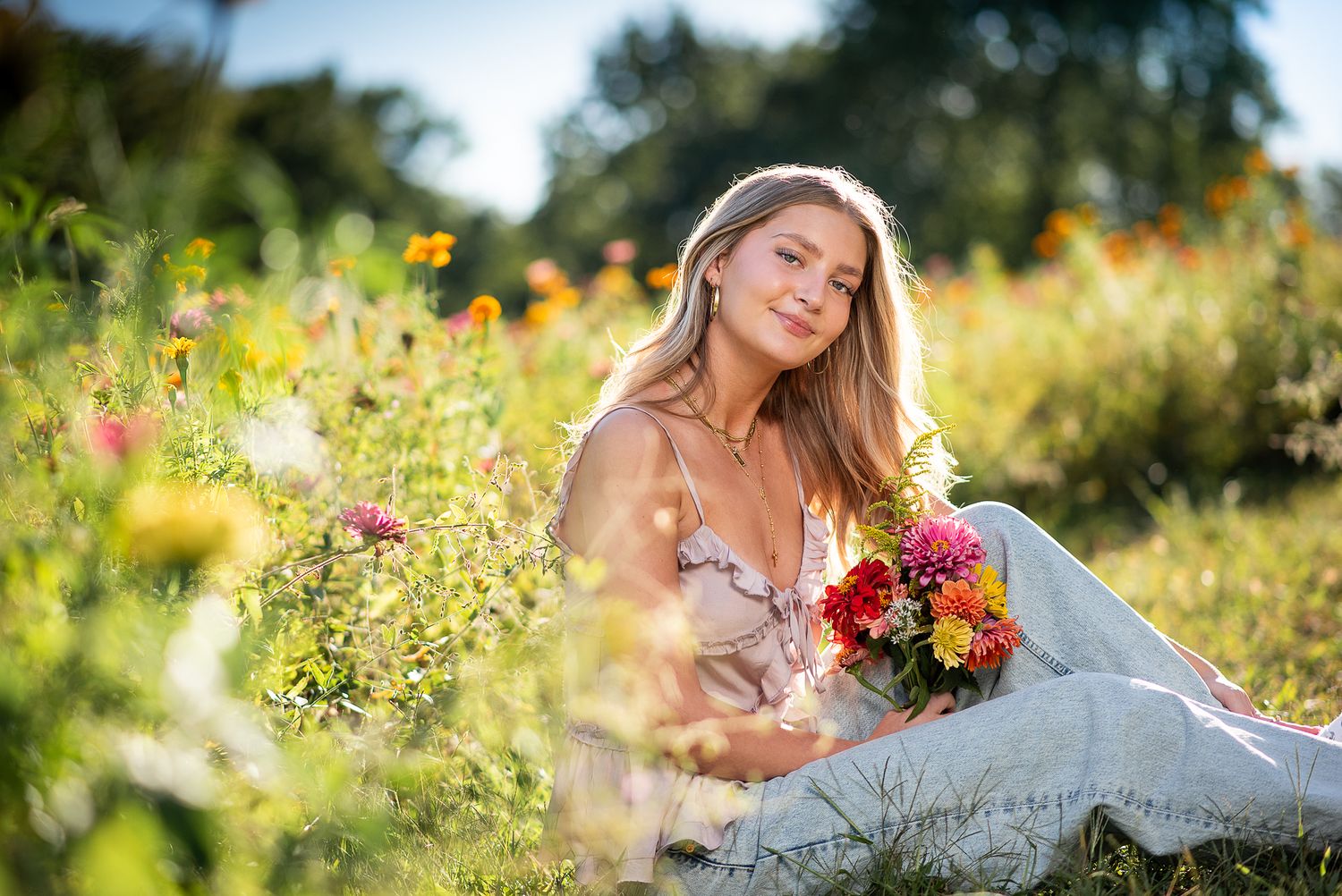 Young woman sitting in a sunny flower garden holding a colorful bouquet, wearing a white outfit.