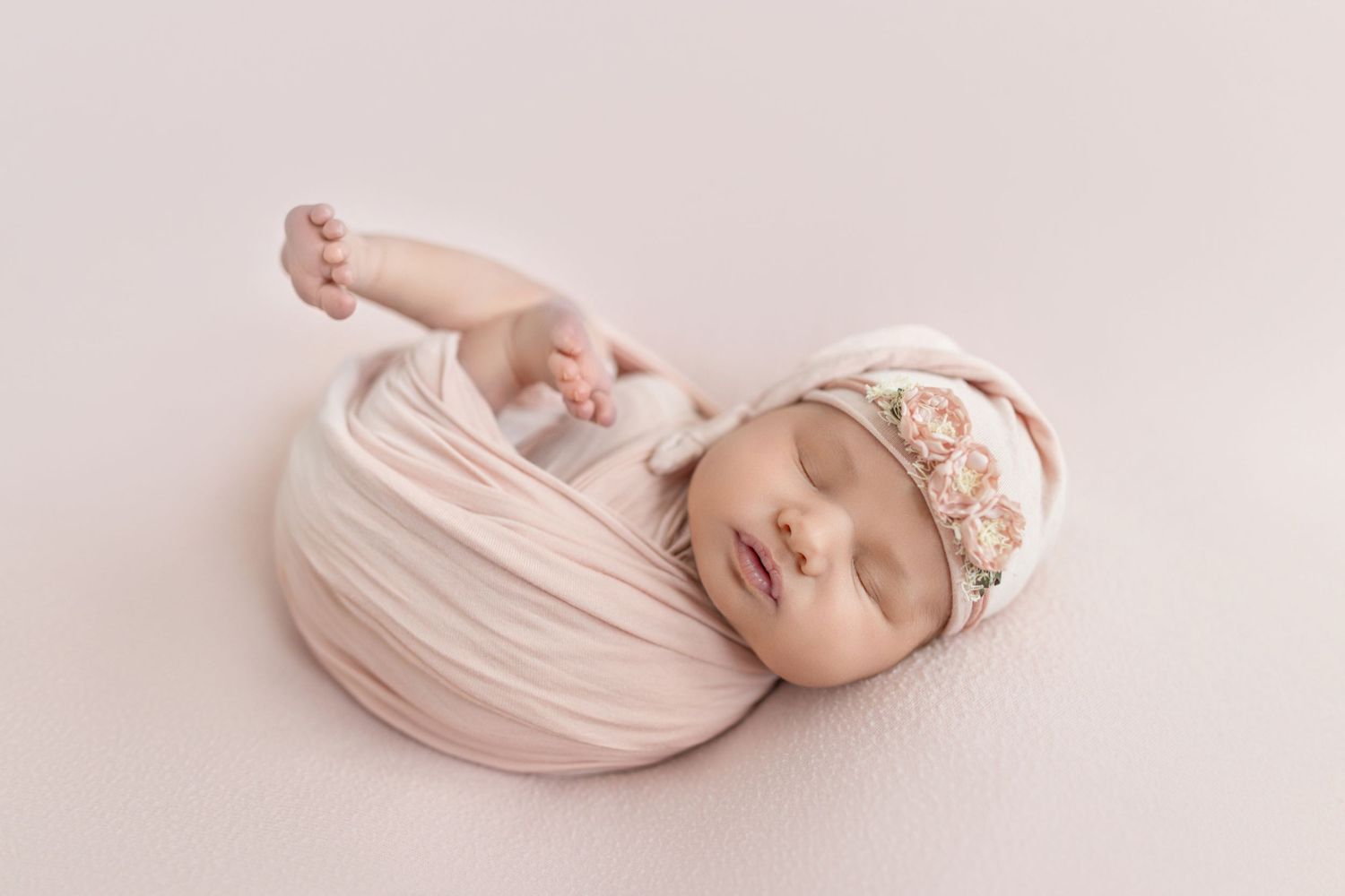 Newborn baby girl wrapped in soft pink swaddle with floral headband, sleeping peacefully in studio photo.