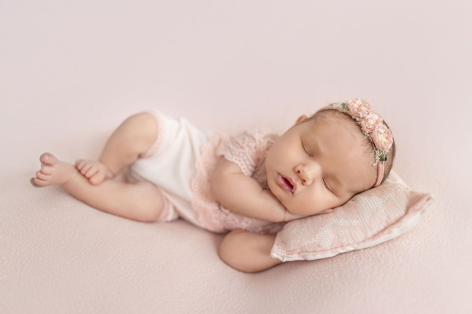 Sleeping newborn baby girl in pink lace outfit resting on a soft pillow with floral headband on pink backdrop.