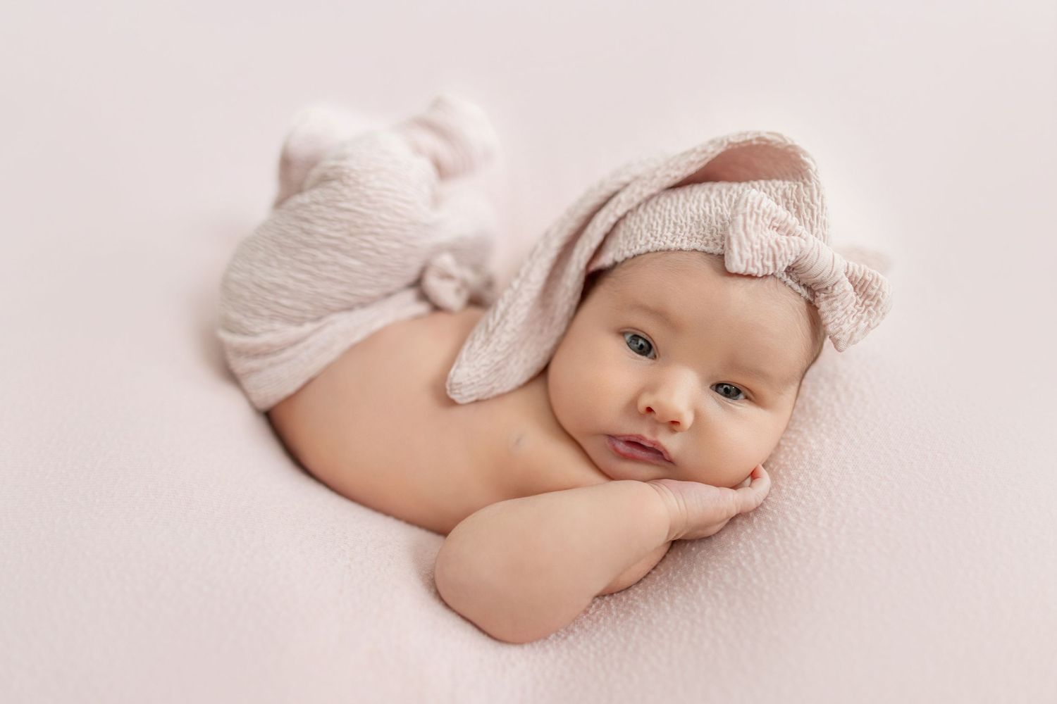 Newborn baby girl lying on a soft white blanket wearing a cute beige bunny ear headband in a professional photo.