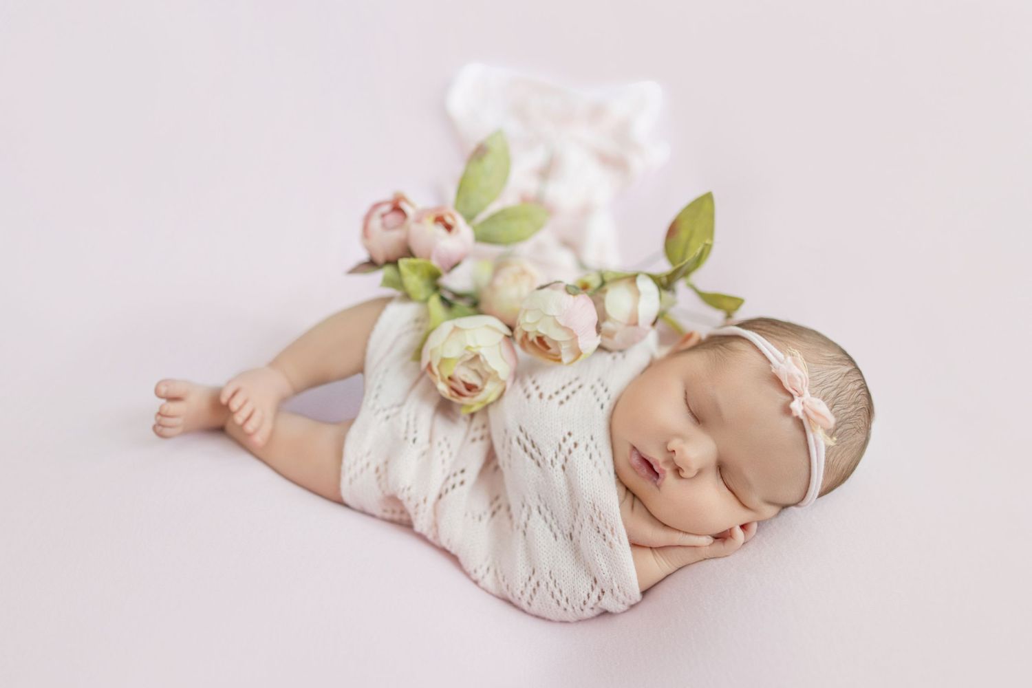 Sleeping newborn baby in white knit romper adorned with soft pink roses on a light pink background.