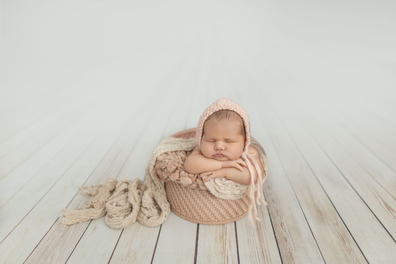 Newborn baby sleeping peacefully in a pink woven basket, wearing a knit bonnet, on a white wood floor.
