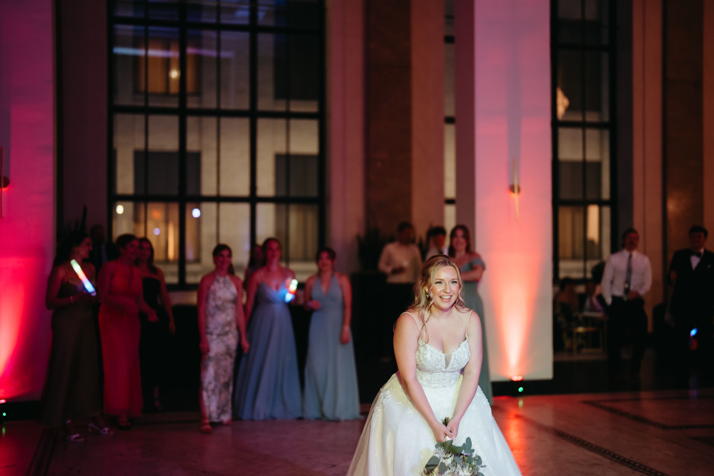 Bride smiling during bouquet toss at elegant wedding reception with pink uplighting and guests in blue gowns.