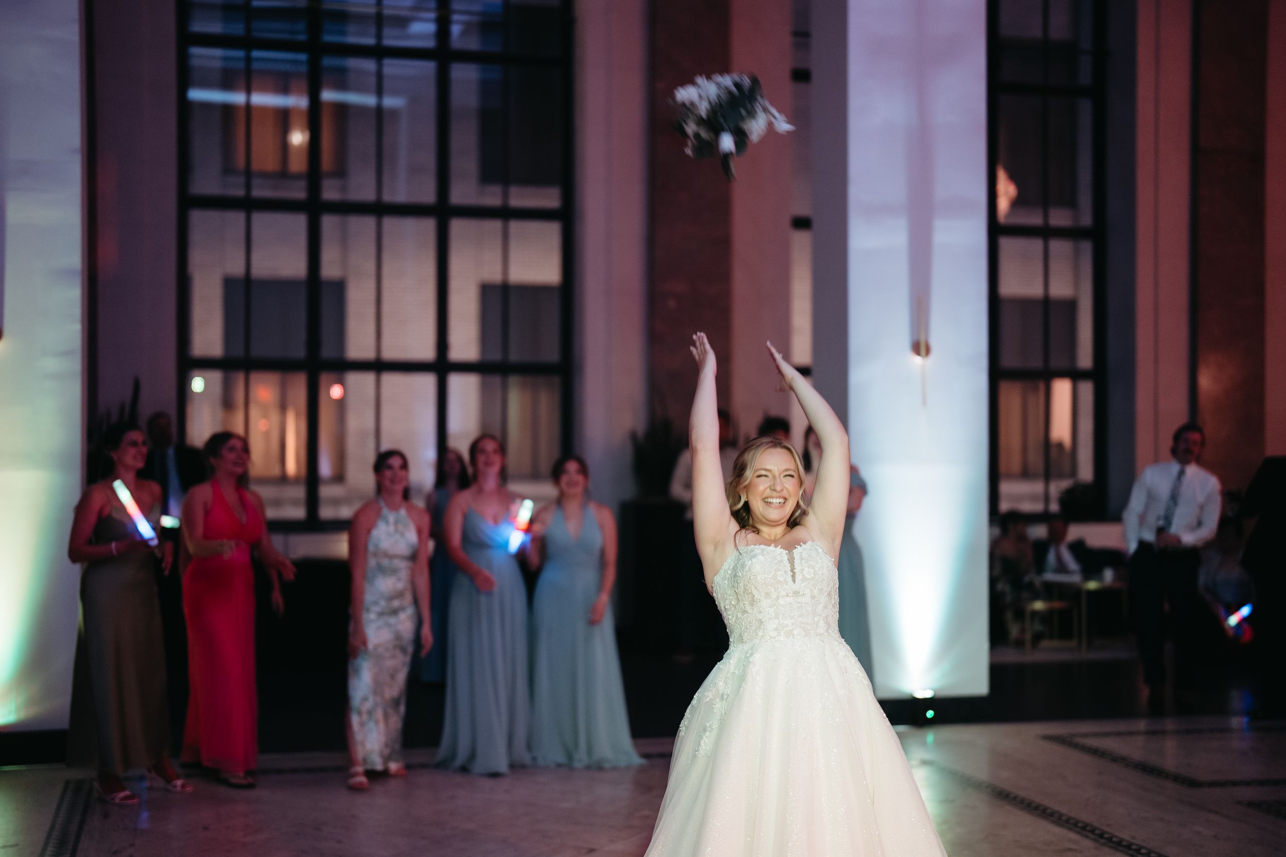 Bride tossing bouquet at wedding reception with guests watching in elegant venue with purple lighting.
