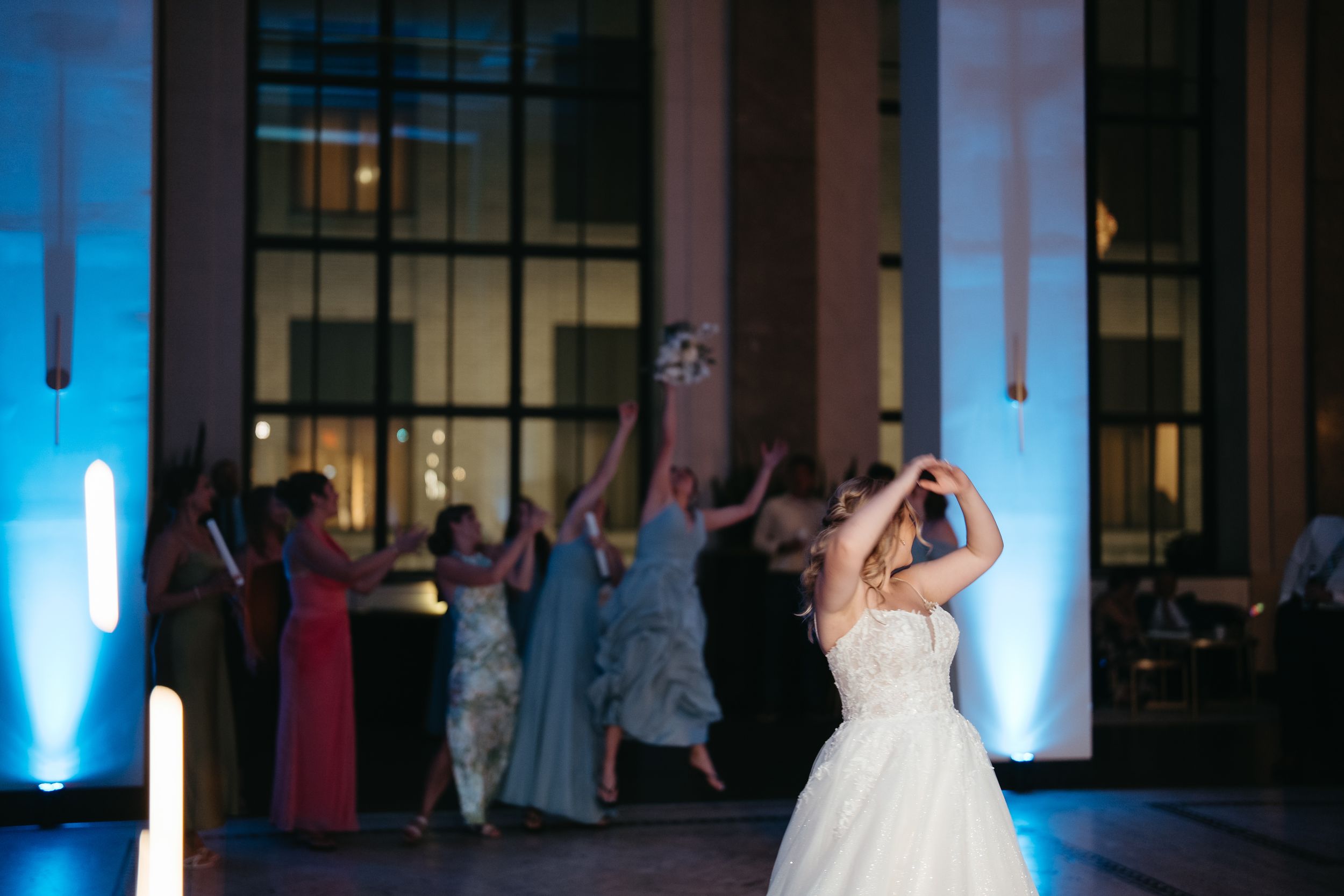 Bride tossing bouquet to excited guests at elegant wedding reception with blue uplighting.