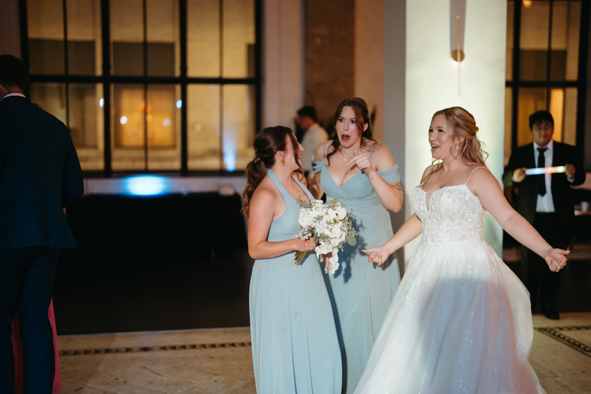 Bride in white gown dancing joyfully with bridesmaids in sage green dresses at elegant wedding reception.
