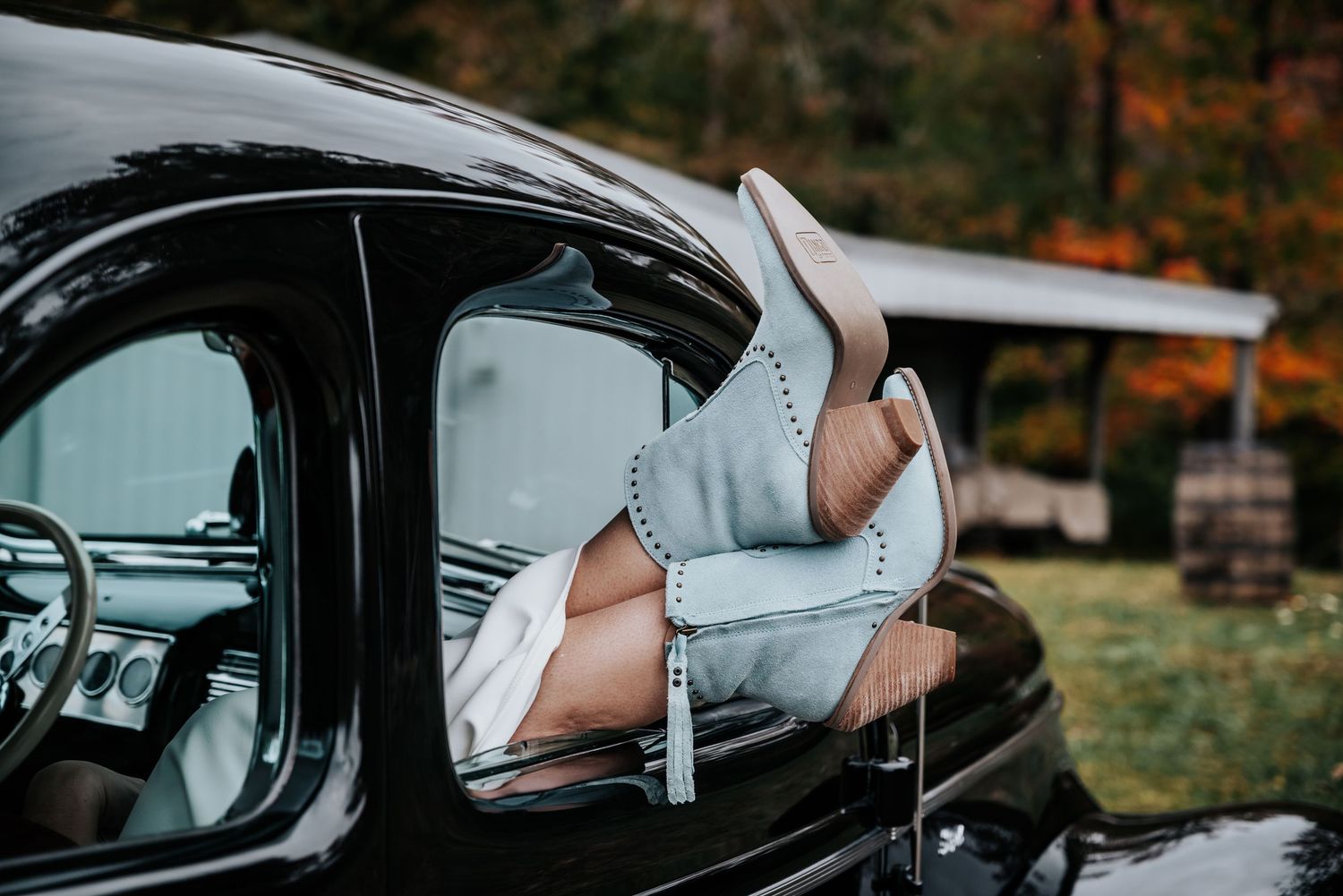 Womens legs in beige heels resting out of a vintage black car window with autumn foliage in background.