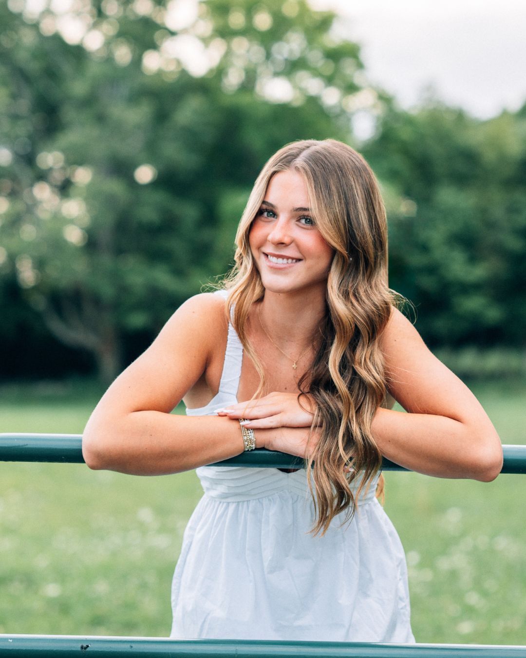 Young woman in white dress leaning on green railing, smiling outdoors with lush green background.