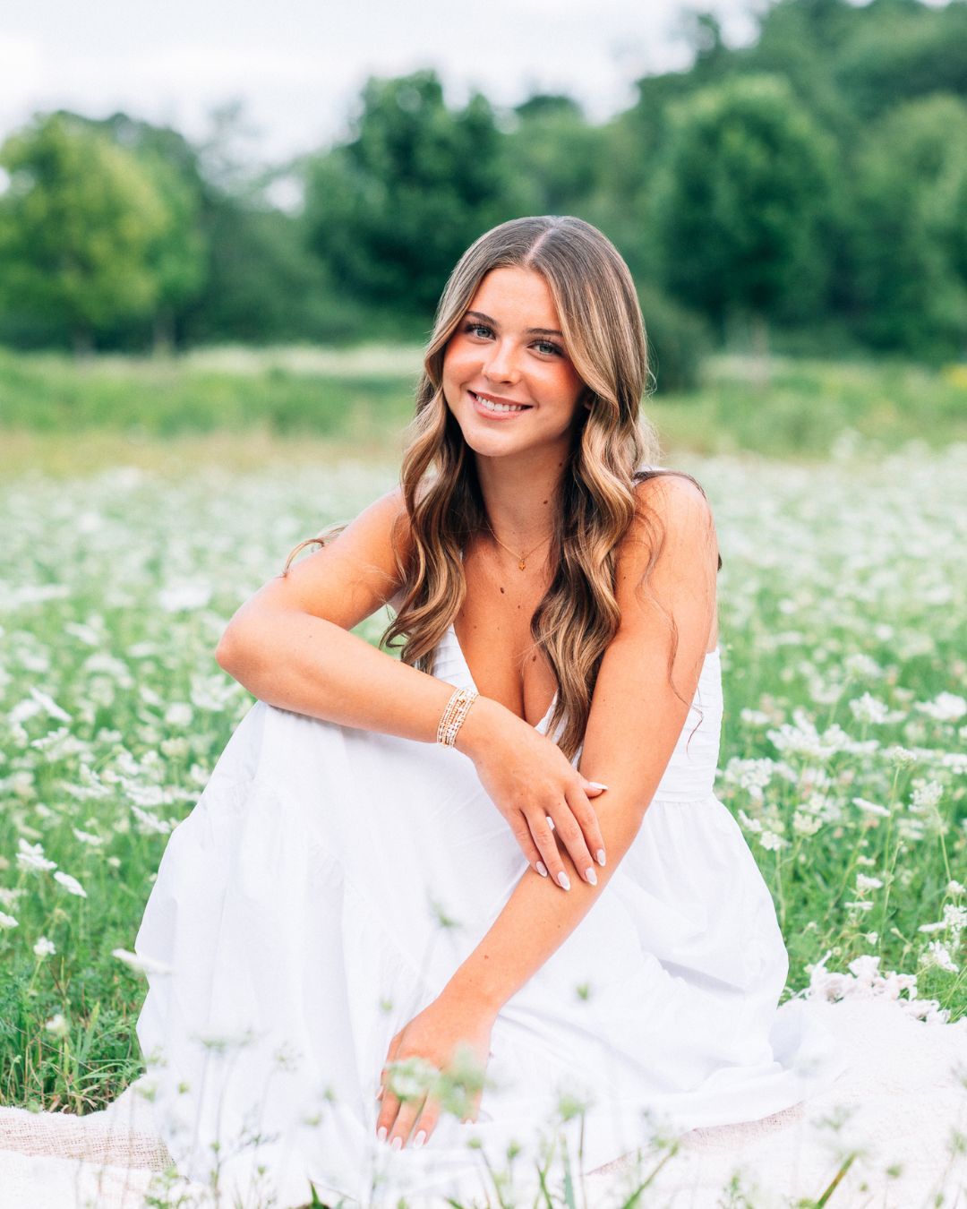 Young woman in white dress sitting in a field of white wildflowers, smiling outdoors on a sunny day.