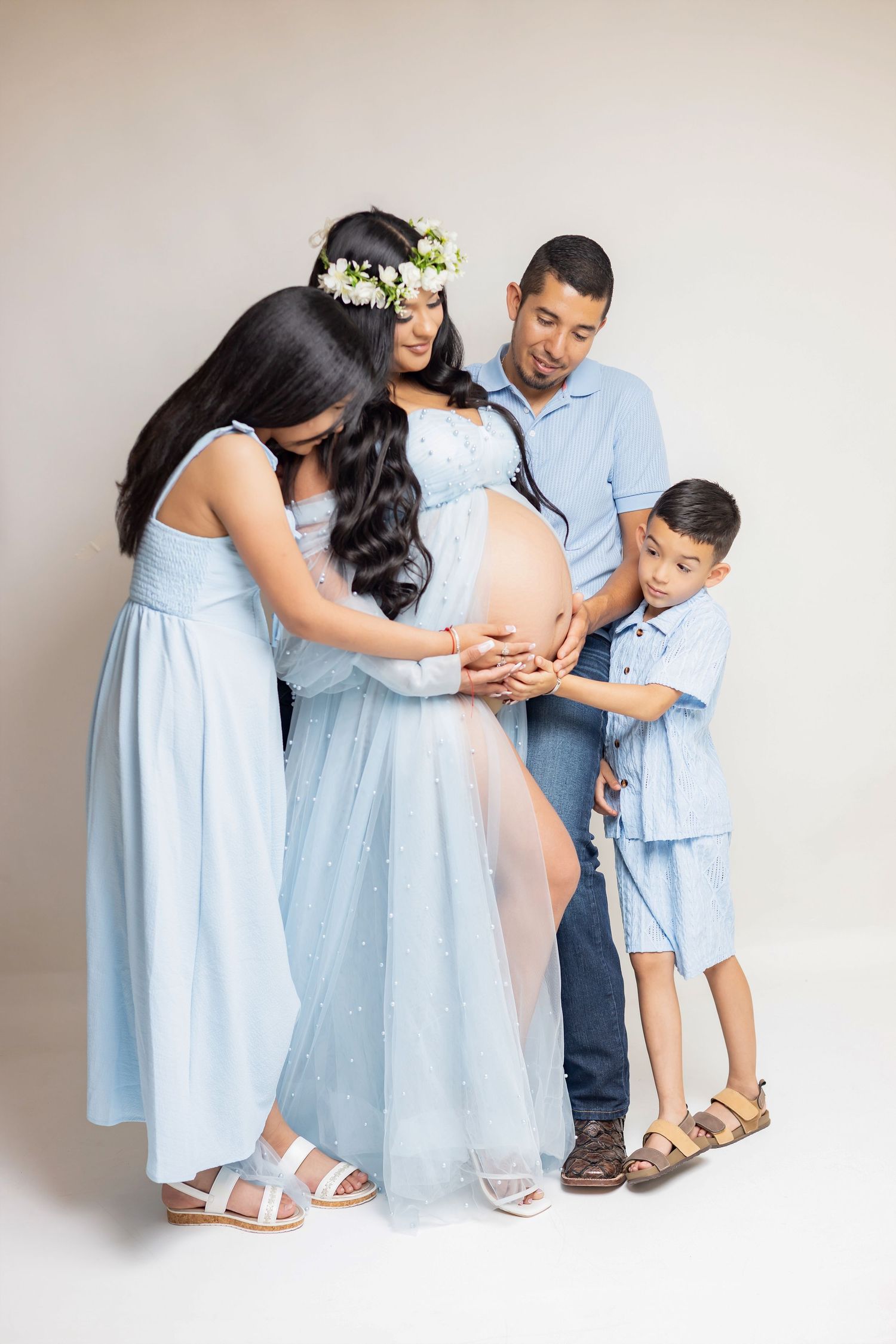 Family maternity photo with pregnant woman in blue gown, flower crown, husband, daughter, and son touching baby bump.