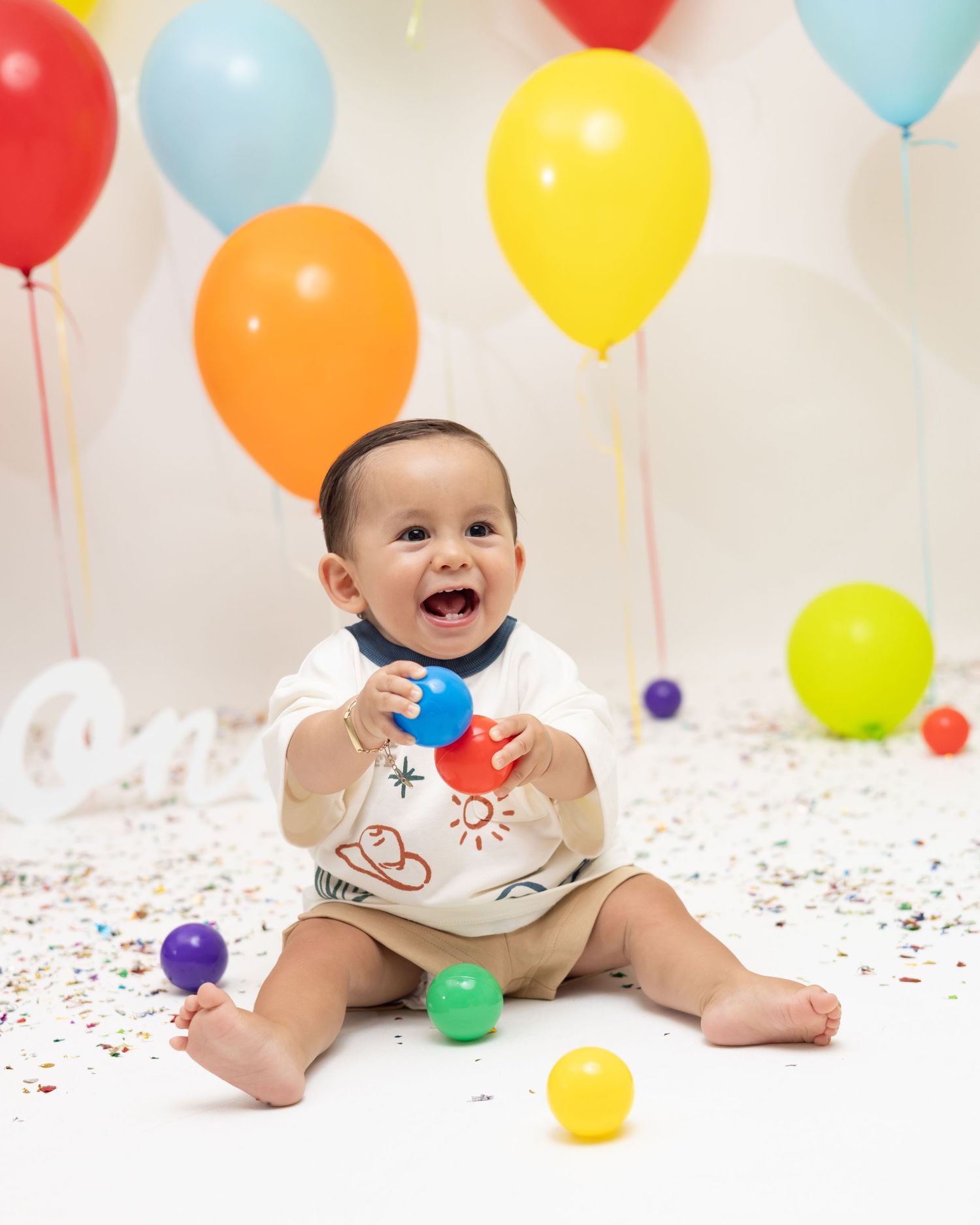 Happy baby sitting on floor surrounded by colorful balloons and confetti, smiling and holding small balls at a first birthday party.