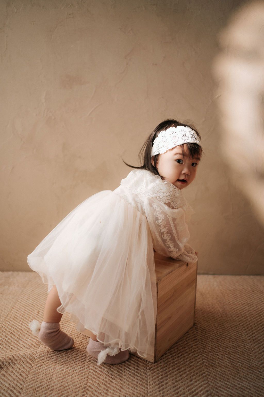 Toddler girl in white tulle dress and lace headband leaning on wooden box in studio photo.