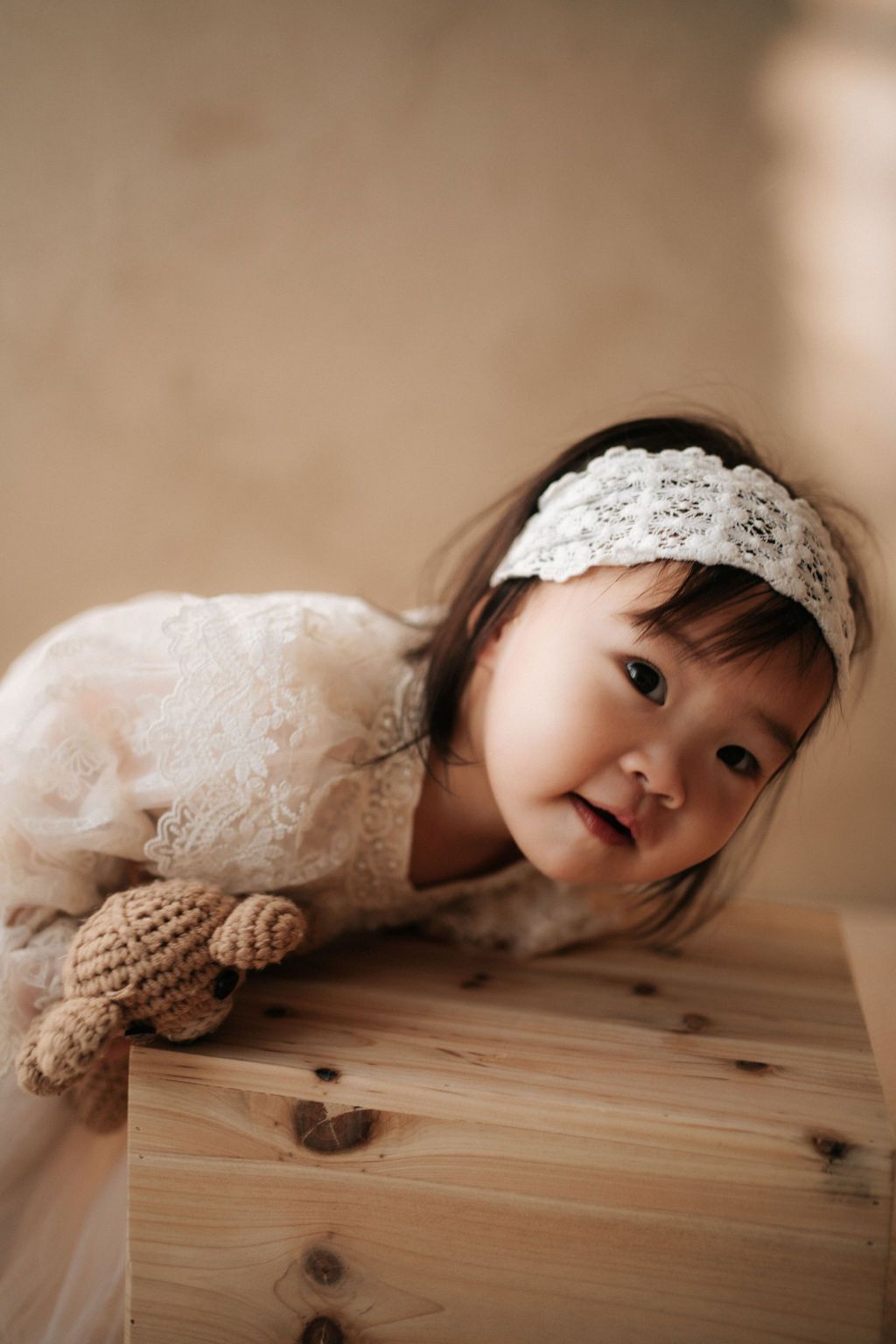 A toddler girl in a white lace dress and headband leans on a wooden box, holding a knit toy.