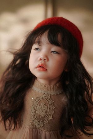 Cute little girl with wavy dark hair and red beret in beige lace dress, gazing upward outdoors.