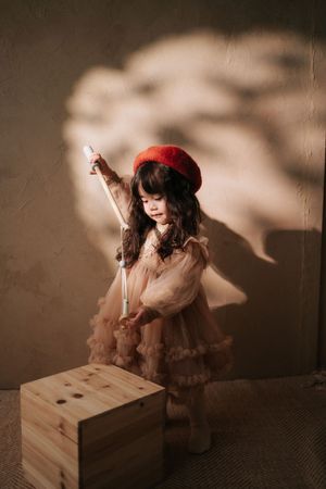 Young girl in red beret and tulle dress holds a white wand beside a wooden cube in warm studio light.