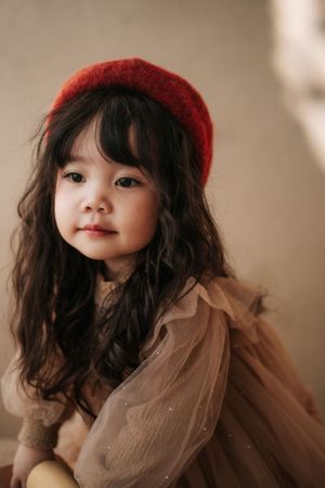 A cute young Asian girl with long dark wavy hair wearing a red beret and beige tulle dress poses softly.