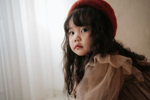 Young girl with curly dark hair wearing a red beret and tulle dress, posing elegantly near a white curtain.