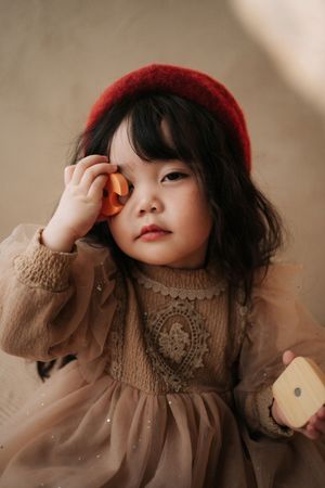 A cute Asian toddler girl in a red beret and beige lace dress holds wooden toys up to her face.