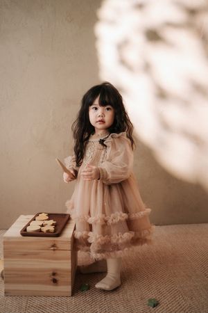 A young girl in a peach tulle dress holds a cookie near a wooden box with heart-shaped cookies on a tray.