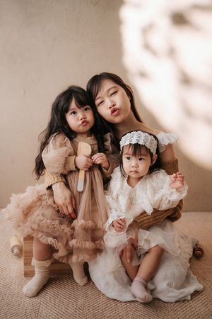 Three young Asian girls in elegant dresses pose together, two making kissy faces in a soft neutral studio setting.