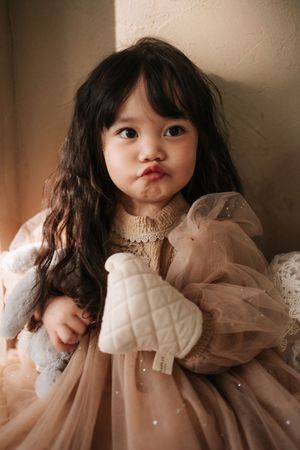 A cute toddler girl with long dark hair wearing a blush pink tulle dress, holding a white quilted toy.