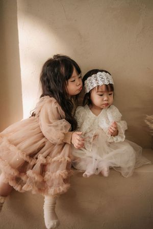 Two young girls in elegant tulle dresses, one kissing the other wearing a lace headband, in a soft beige setting.
