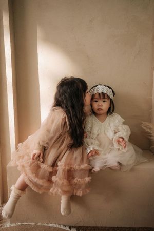 Two young Asian girls in elegant dresses, one whispering to the other, sitting in soft natural light.