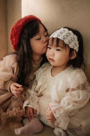 Two young Asian girls in vintage lace dresses, older sister in red beret kissing younger toddler's cheek.