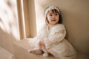 Adorable toddler girl in white lace dress and headband sitting by a window in soft natural light.