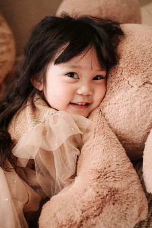 Smiling toddler girl with dark hair in a beige tulle dress cuddling a large fluffy teddy bear.