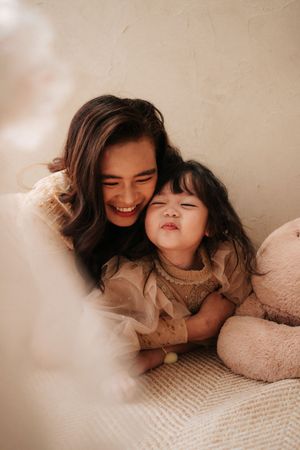 A smiling woman cuddles a laughing toddler girl in matching beige outfits on a cozy textured surface.