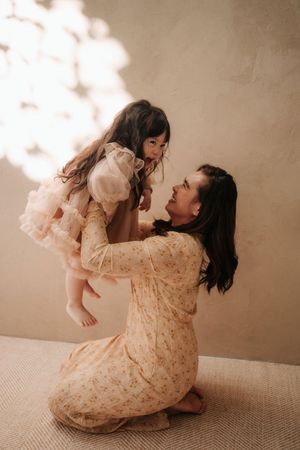 A mother in a floral dress kneels while lifting her young daughter in a tulle dress, sharing a joyful moment.