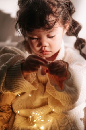 A young girl in a white knit sweater plays with glowing fairy lights, her expression focused and curious.