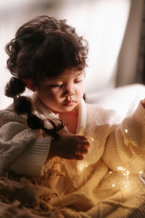 Toddler girl with curly pigtails in white knit sweater playing with fairy lights on cozy blanket.