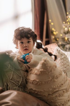 Cute toddler girl with dark curly pigtails in white sweater holding blue ornament near Christmas tree.