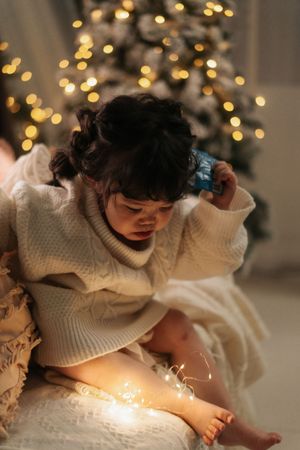 A young child in a white knit sweater examines a small blue gift near a glowing Christmas tree with fairy lights.