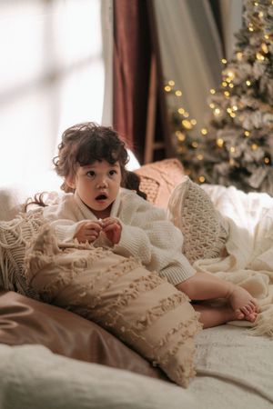 Toddler in cream knit sweater sitting on bed with pillows near a glowing Christmas tree.