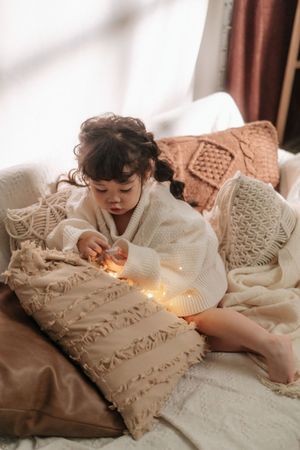 Toddler girl in white knit sweater playing with fairy lights on cozy boho pillows and blankets on a sofa.
