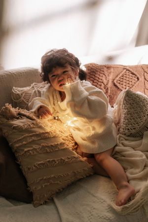 Toddler in white knit sweater sitting among cozy pillows holding glowing fairy lights in warm light.
