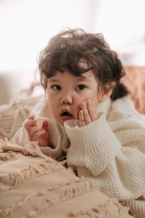 Adorable toddler in a white knit sweater lying on a beige fringed blanket, looking curiously at the camera.