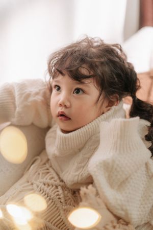 A cute toddler with curly dark hair wearing a cozy cream knit turtleneck sweater surrounded by warm bokeh lights.