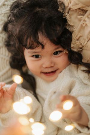 A smiling toddler with dark curly hair lies on a cozy blanket, reaching toward glowing fairy lights.