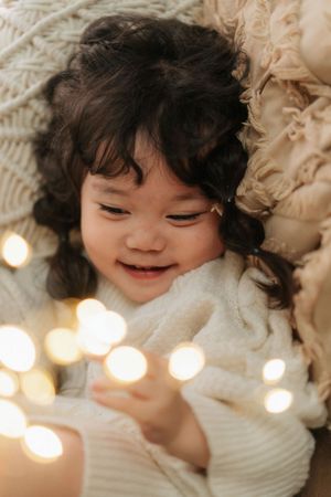 Smiling toddler with dark curly hair lying among warm bokeh fairy lights in a cozy cream knit sweater.