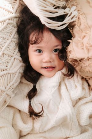 Adorable toddler girl with dark pigtails wearing a white cable-knit sweater, nestled among cozy cream blankets.