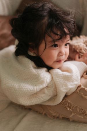 Adorable Asian toddler girl in a white knit sweater lying on a couch, smiling softly with dark curly hair.