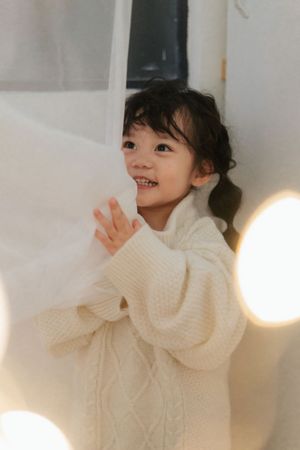A smiling young Asian girl in a cream cable-knit sweater holds a white curtain, surrounded by soft bokeh lights.